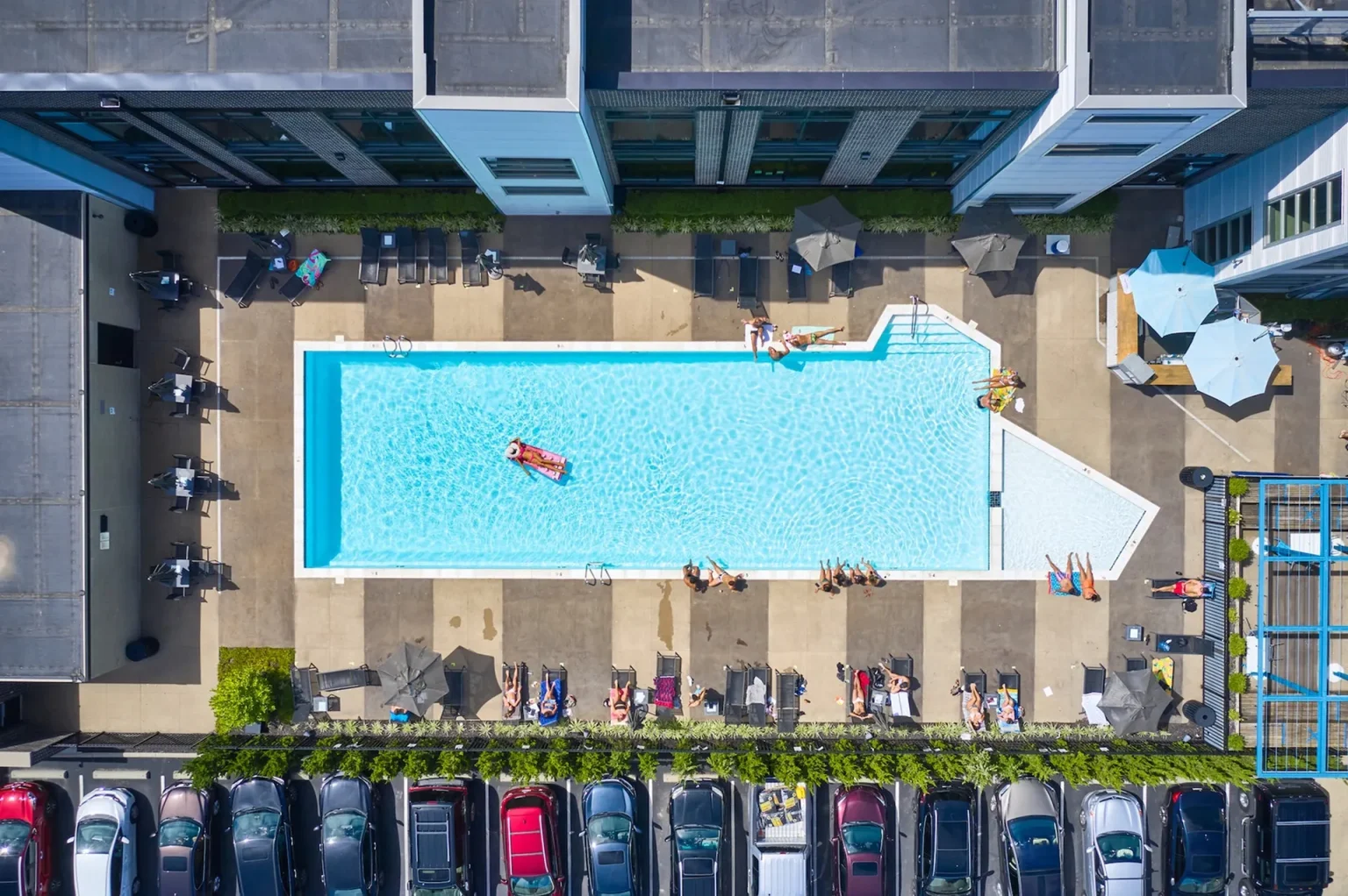 Aerial view of a rectangular outdoor pool at Columbus apartments, surrounded by lounge chairs and umbrellas. Residents bask in the sun while others enjoy a refreshing swim. Nearby, a row of parked cars hints at the convenience this vibrant community offers.