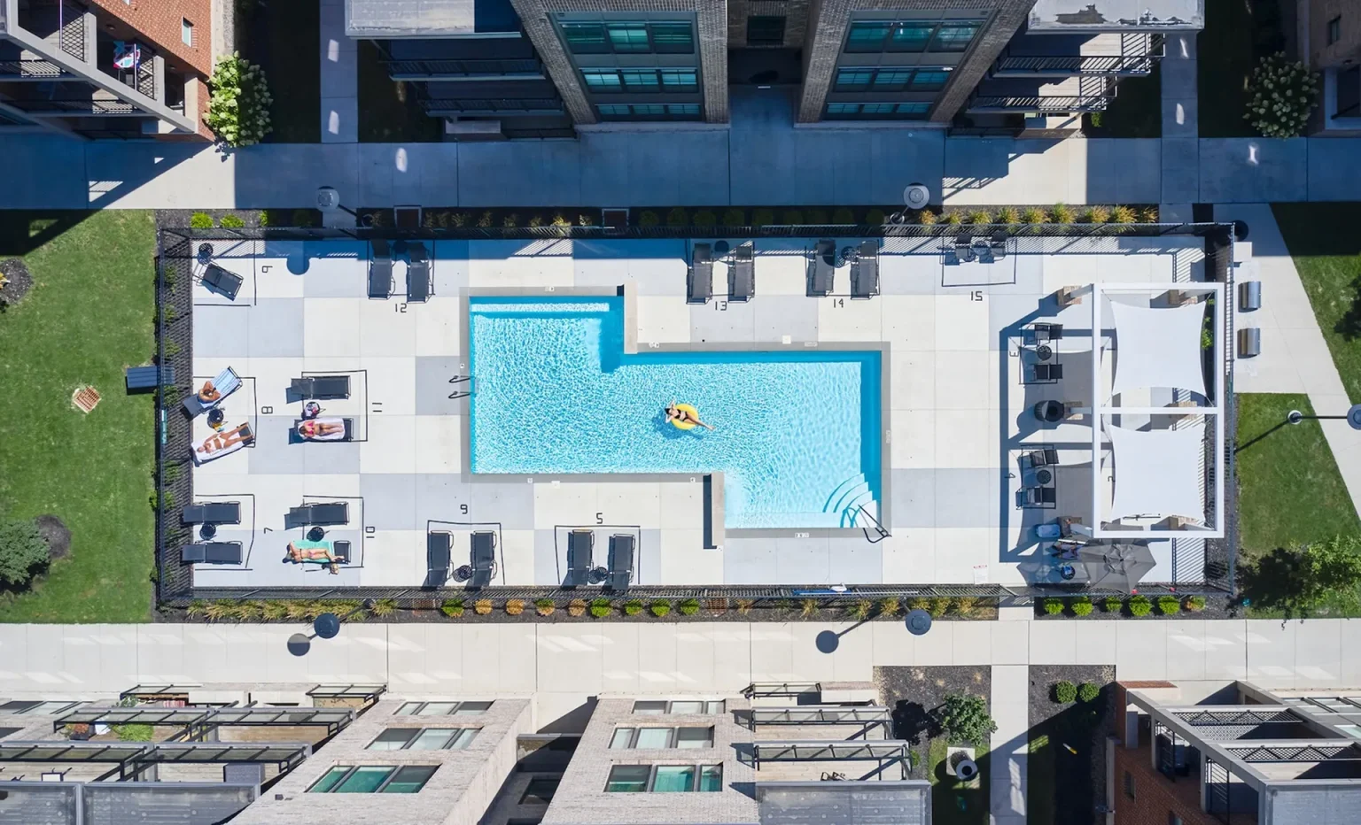 An aerial view of the Columbus Apartments reveals a rectangular outdoor swimming pool surrounded by lounge chairs and buildings. A person is leisurely floating on a yellow inflatable in the pool, enjoying the serene ambiance of this charming residential oasis.
