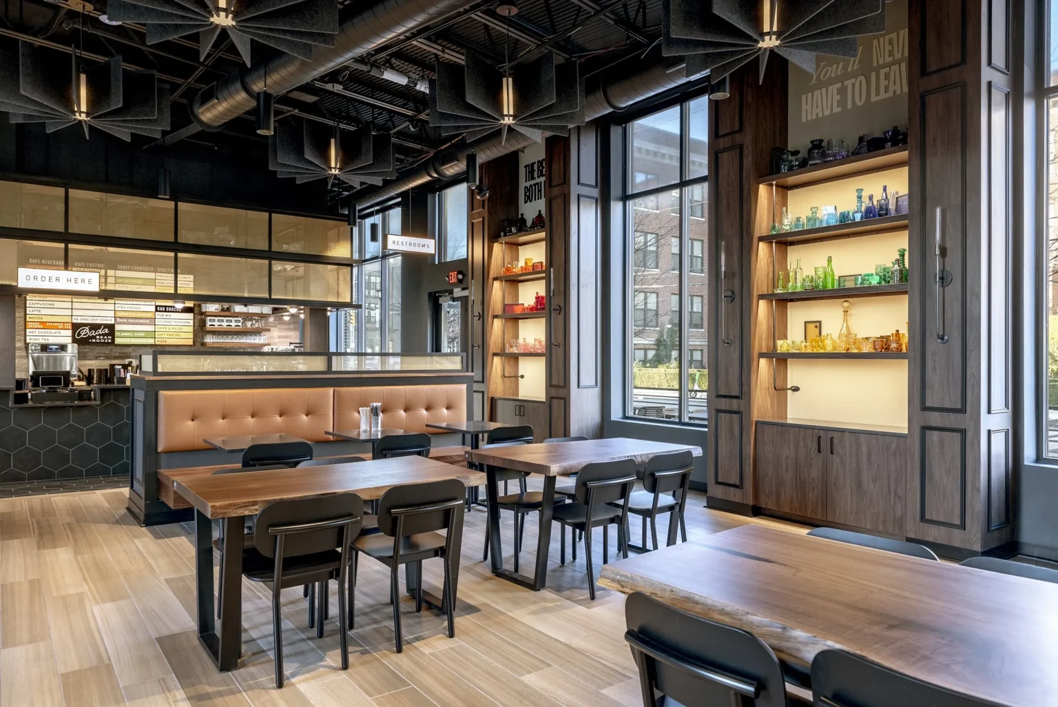Modern cafe interior reminiscent of stylish Columbus apartments, featuring wooden tables, black chairs, and a padded bench. Colorful decorative bottles adorn the shelves, while large windows provide natural light. An inviting order counter is stationed in the background.