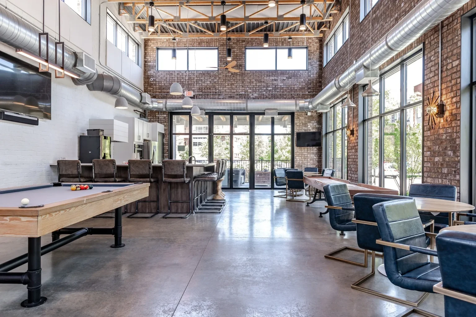 A spacious room in Columbus apartments features a pool table, bar area, and seating with tables and chairs. Large windows invite natural light, while exposed ductwork and brick walls add an industrial charm.