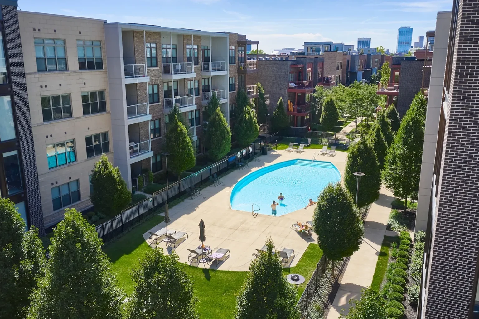 Columbus apartments offer a serene escape with a central outdoor pool, surrounded by trees and pathways. Residents enjoy swimming and lounging on deck chairs under a clear blue sky, while the vibrant urban skyline provides a stunning backdrop.