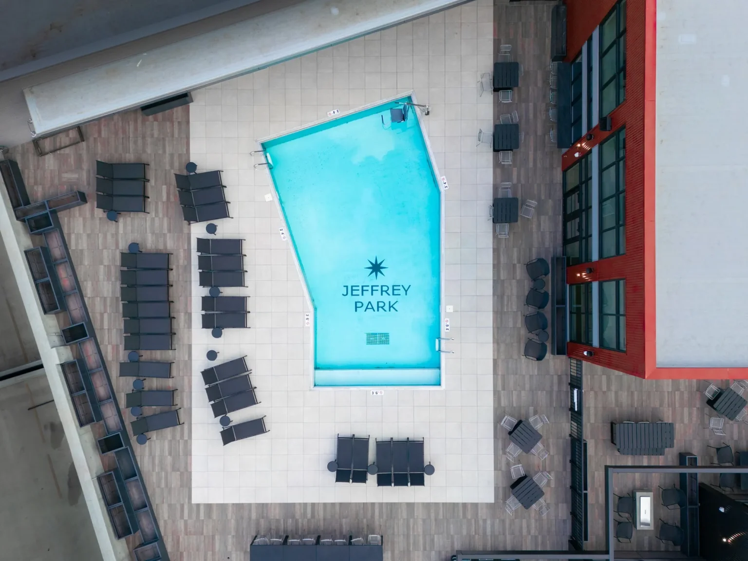 Aerial view of a rectangular swimming pool labeled Jeffrey Park, nestled within the vibrant Columbus apartments, surrounded by lounge chairs and tables on a concrete patio adjacent to a red building.