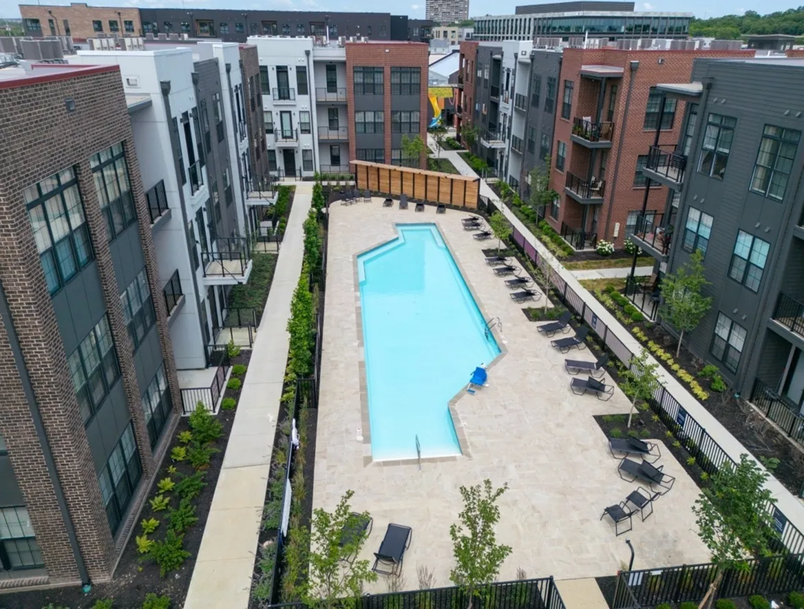 Aerial view of Columbus apartments reveals a residential complex with a central outdoor pool, surrounded by lounge chairs and lush greenery. Multiple multi-story buildings are harmoniously nestled within the vibrant community landscape.