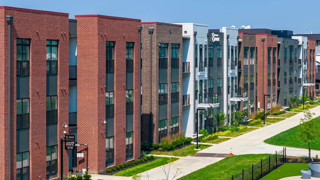 Row of modern, multi-story Columbus apartments with brick and glass exteriors, adjacent to a sidewalk and landscaped greenery under a clear blue sky.