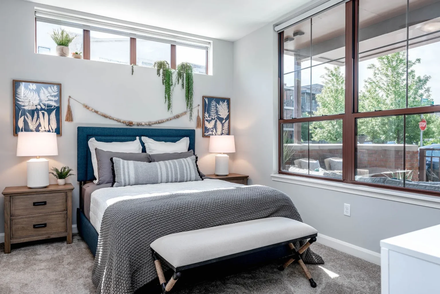 A bedroom in Columbus apartments boasts a bed with gray and blue bedding, two wooden nightstands with lamps, a bench at the foot, and large windows letting in natural light. Wall decor includes art and greenery.