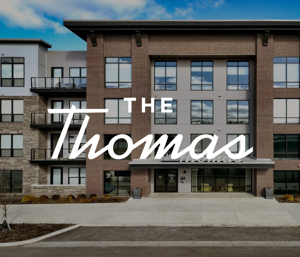 Front view of a modern apartment building with brick and glass facade. Large text overlay reads The Thomas. Discover sophisticated Columbus apartments offering urban elegance and style.