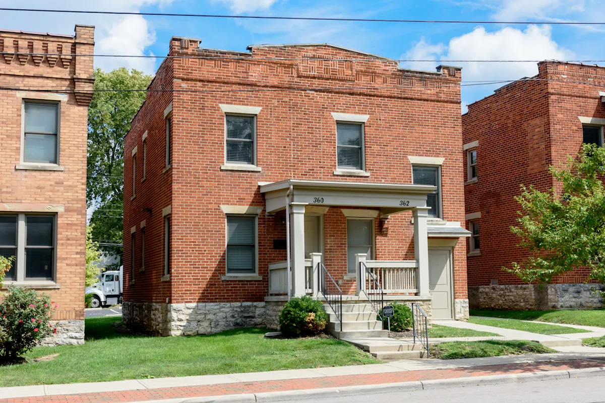 Two-story red brick building with white-trimmed windows and entrance. A small front porch with steps leads to the door. Lawn in front, neighboring buildings on either side, and trees behind.