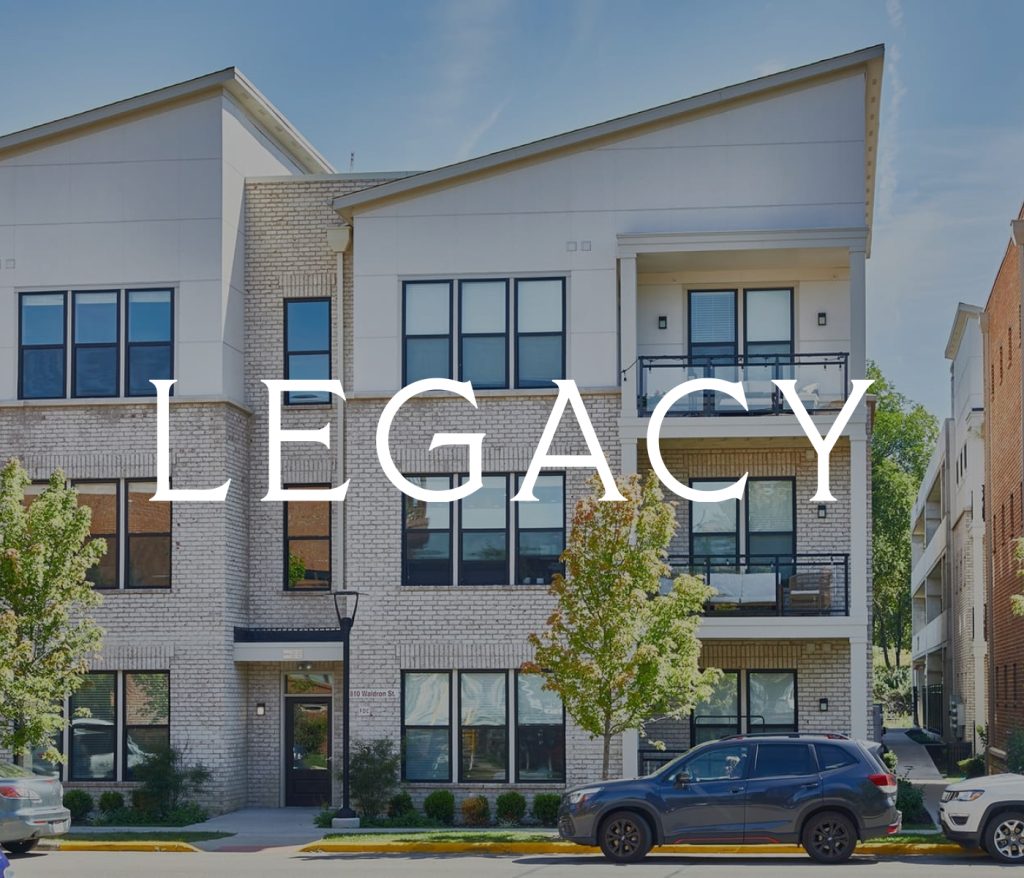 Three-story modern apartment building with Legacy text overlay, exemplifying the charm of Columbus apartments. Cars are parked on the street, and lush trees stand gracefully in front.