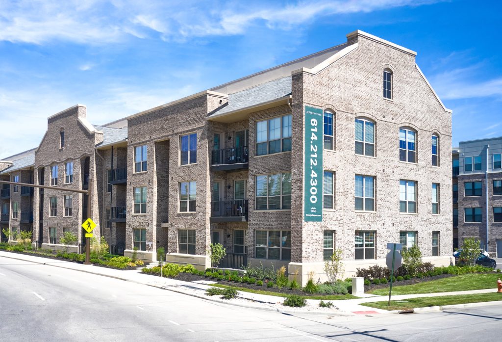 The three-story brick Columbus apartment building boasts charming balconies and is conveniently adjacent to a street. A large vertical sign highlights a phone number, all under a clear blue sky.
