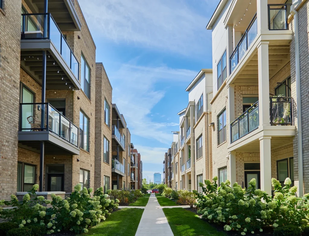 Pathway between two rows of modern apartment buildings with balconies, surrounded by landscaped greenery under a clear blue sky.