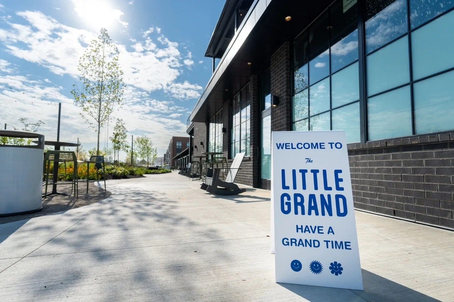 A sandwich board sign graces the sunny sidewalk, cheerfully announcing, Welcome to The Little Grand. Have a grand time, accompanied by smiley, flash, and snowflake icons. Nearby, modern Columbus apartments rise against a backdrop of trees.