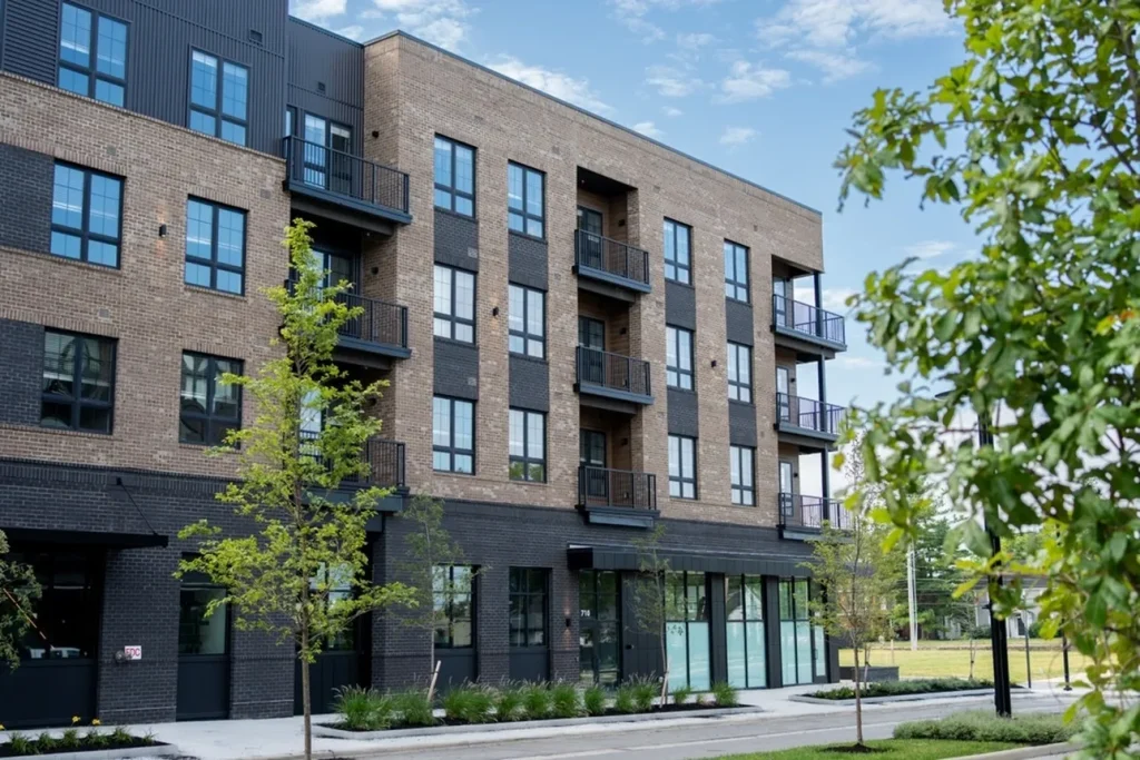 Four-story brick apartment building with large windows and balconies, surrounded by young trees and greenery, under a clear blue sky.