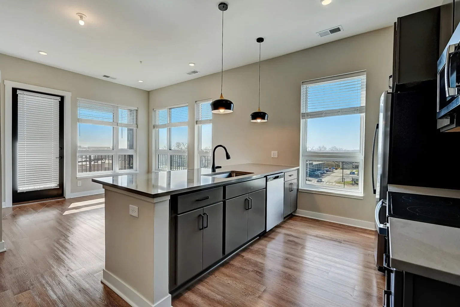 Modern kitchen in Columbus apartments features a large island, black cabinets, and stainless steel appliances. Pendant lights hang elegantly above the island. Large windows offer natural light and scenic views, with wood flooring throughout enhancing the sleek design.