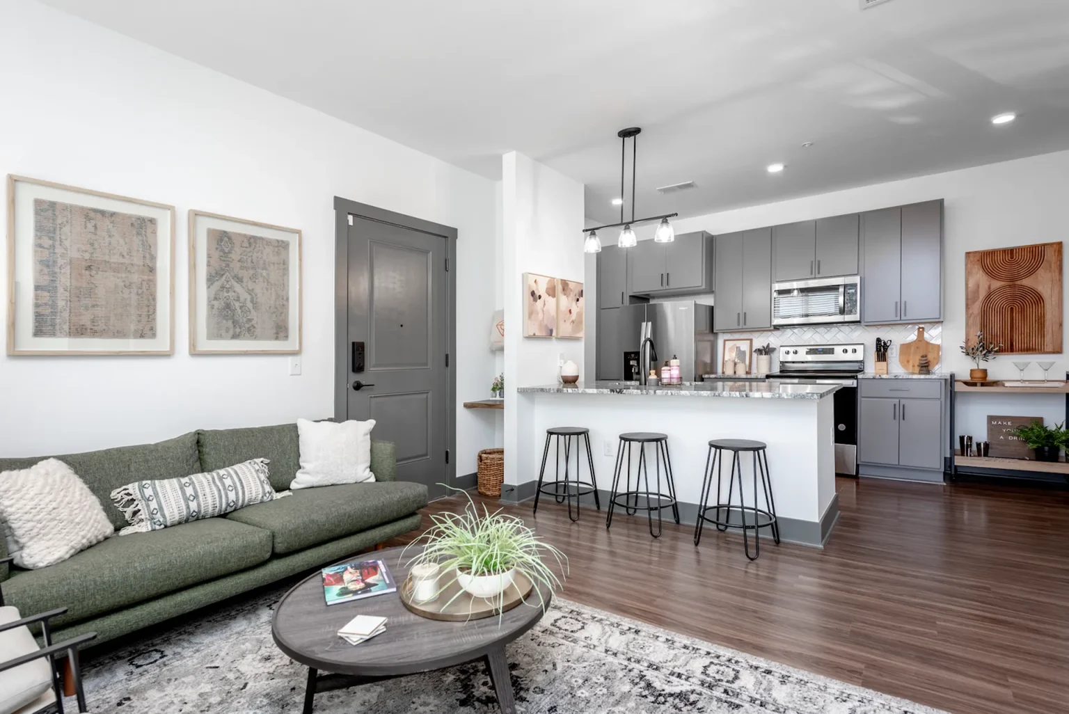 Modern open-concept living room and kitchen in Columbus apartments, featuring a green sofa, round coffee table, bar stools at a white kitchen island, and wall art. Wood flooring and contemporary decor complete the stylish space.