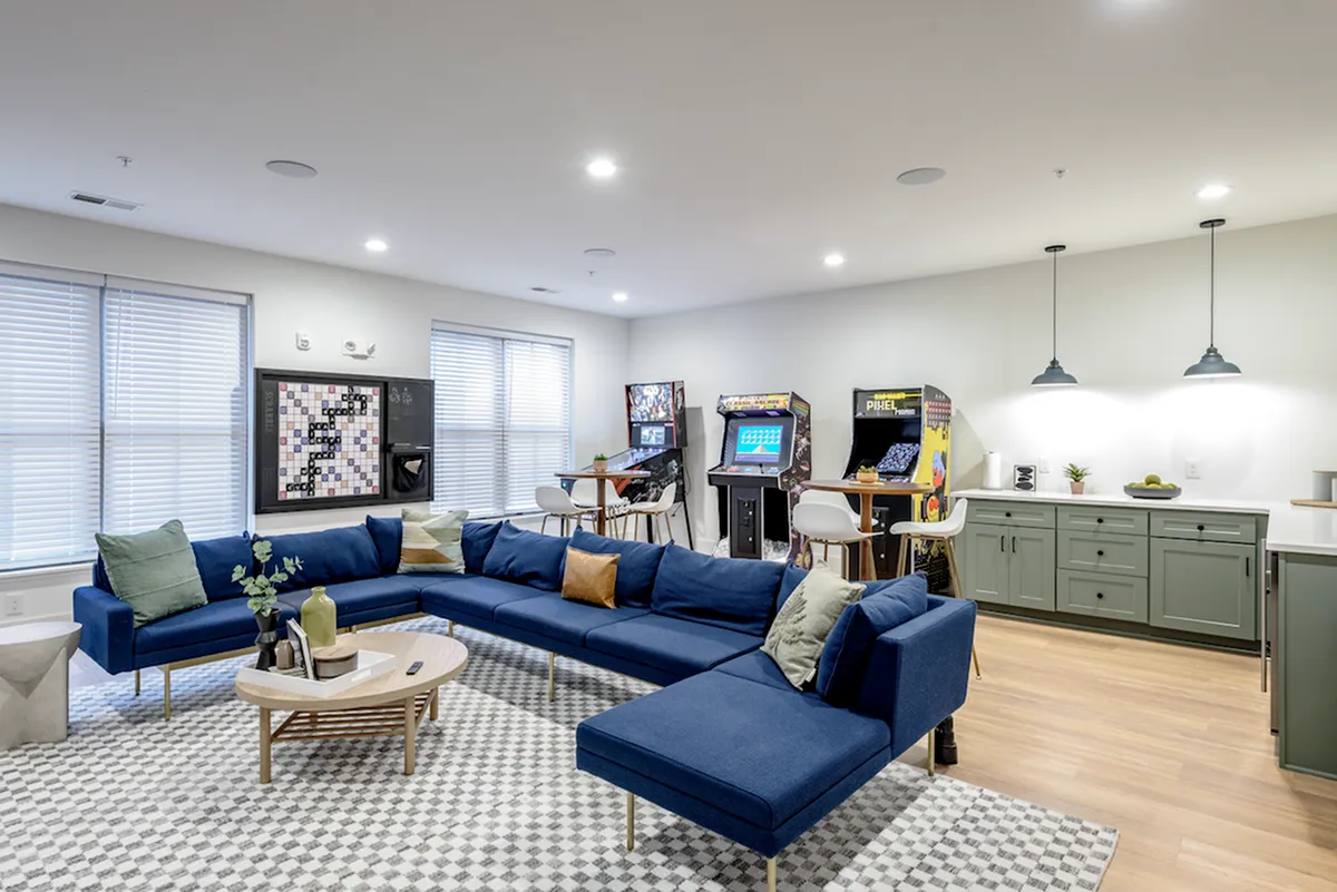 Experience the charm of Columbus apartments in this modern recreational room featuring a blue sectional sofa, arcade machines, and vibrant wall art. Green cabinets add style while large windows provide ample natural light.
