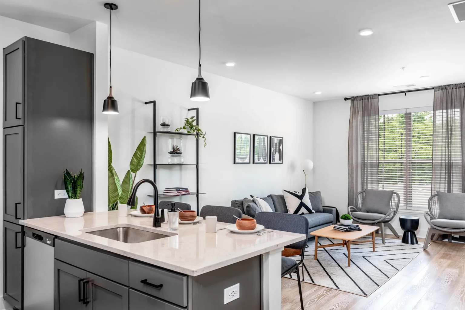 Located in Columbus, this modern open-concept living room and kitchen boasts a kitchen island with a sink, complemented by a gray sofa, armchairs, wall art, and lush plants. Bright and airy with large windows and neutral decor, the space exudes comfort and style typical of Columbus apartments.