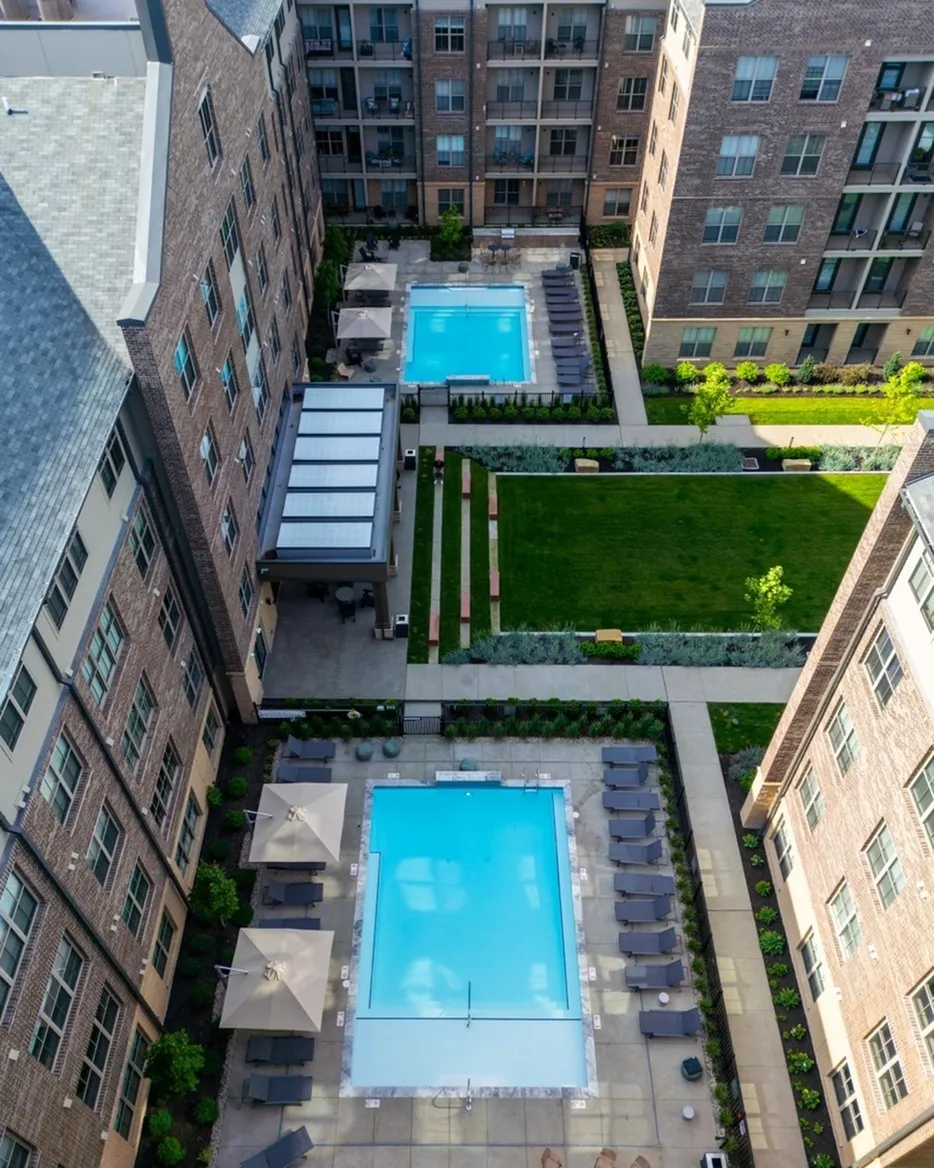 Aerial view of Columbus apartments showcases a large rectangular courtyard with two shimmering swimming pools, surrounded by brick buildings and lush green lawn areas offering inviting seating spaces.