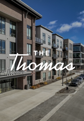 Modern apartment building exterior with The Thomas written in large white script across the center of the image. Cars are parked along the street in front, showcasing stylish Columbus apartments in a vibrant urban setting.