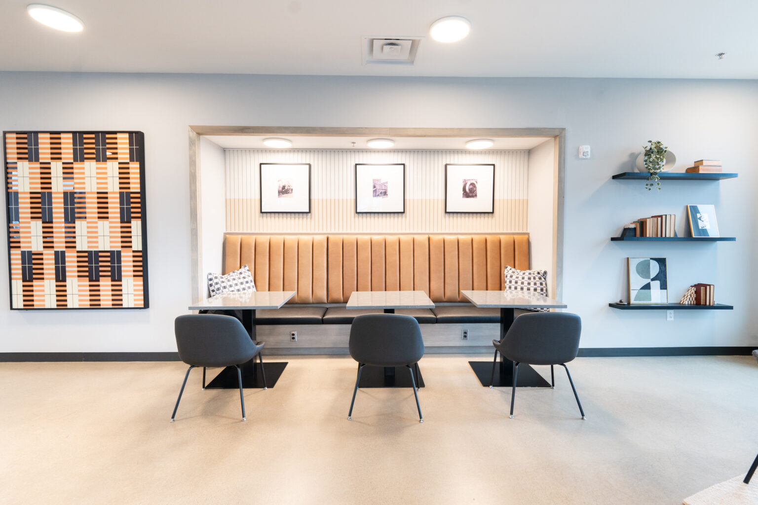 Modern seating area in Columbus apartments with a cushioned booth, four chairs, two tables, wall art, and shelves with books and decor. Bright lighting and neutral colors create a clean, minimalist look.