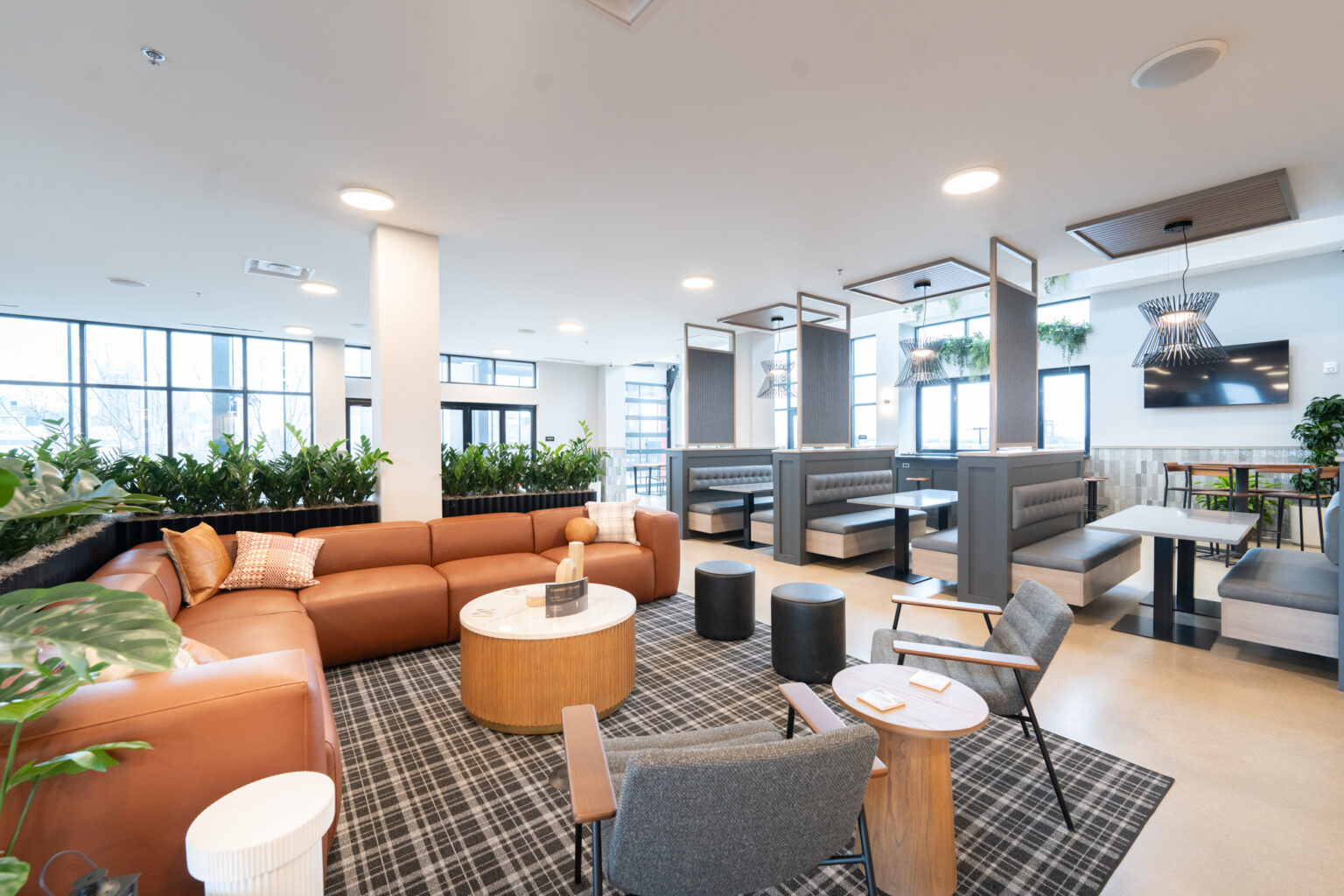 Modern lounge area in Columbus apartments featuring a brown sectional, round tables, and chairs on a checkered rug. Booth seating with wooden tables is in the background. Green plants and large windows provide natural light.