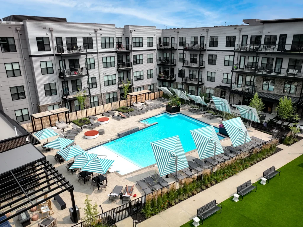 Aerial view of a modern apartment complex with a central outdoor pool, surrounded by lounge chairs and striped umbrellas, adjacent to a grassy area with benches.