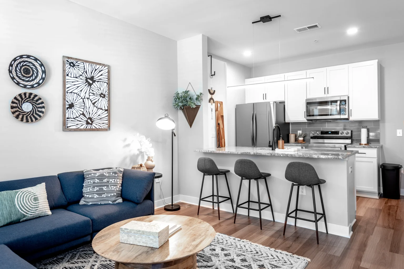 Modern open-concept living room and kitchen in Columbus apartments, featuring a blue couch, wooden coffee table, three gray bar stools, and stainless steel appliances. Decor includes wall art and patterned cushions.