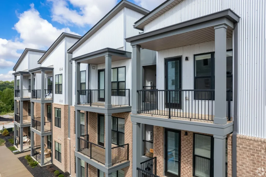 Modern apartment building with three floors, featuring balconies and a mix of brick and white panel exterior. The sky is partly cloudy in the background.
