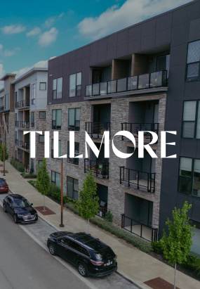 Modern apartment building with balconies, stone and wood exterior, parked cars, and tree-lined street; TILLMORE AT QUARRY TRAILS text overlay in bold white font.