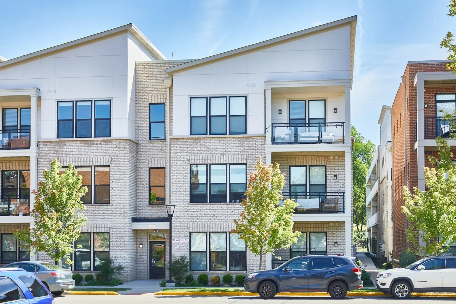 This modern three-story Columbus apartment building boasts a brick facade, large windows, and inviting balconies. Two parked cars and small trees enhance its charm on a sunny day.