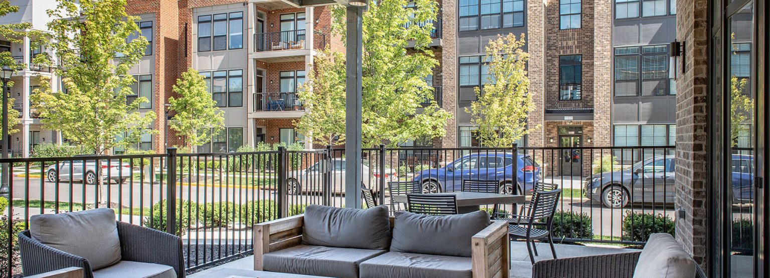 Outdoor patio with gray cushioned chairs and a small table overlooks a street with parked cars and modern apartment buildings in the background.