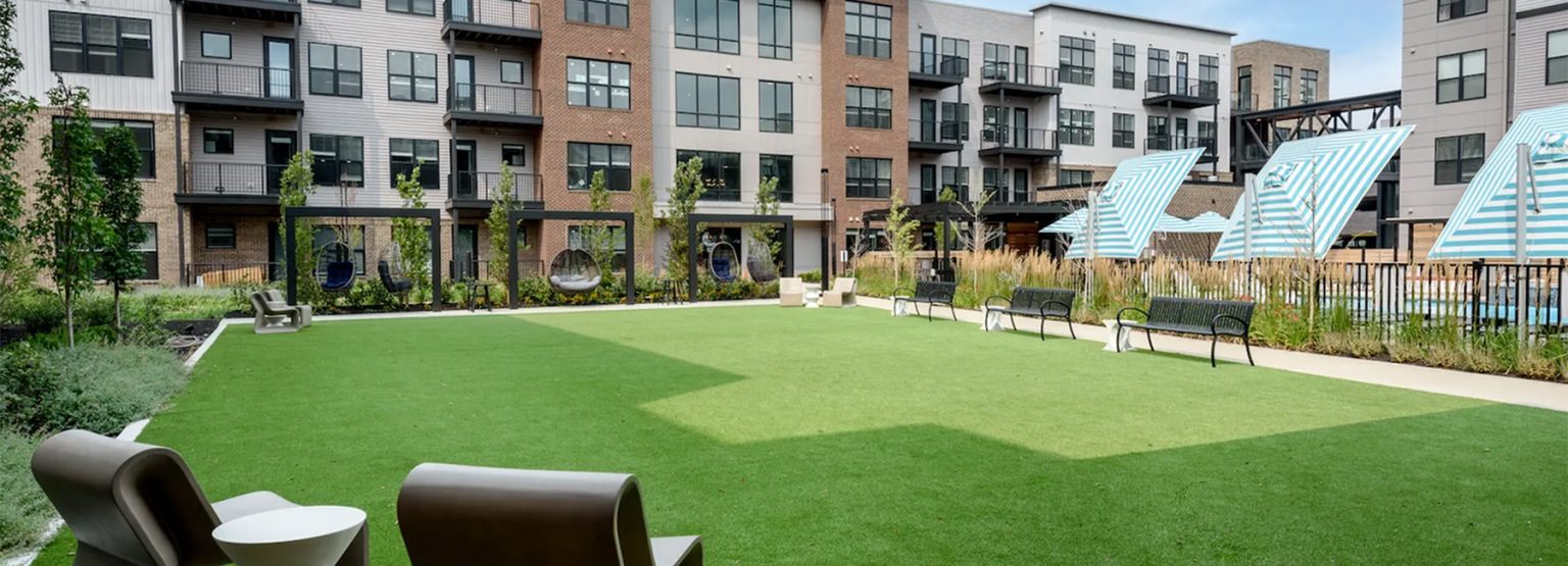 Courtyard with green artificial lawn, chairs, benches, and modern apartment buildings. Striped cabanas are in the background.