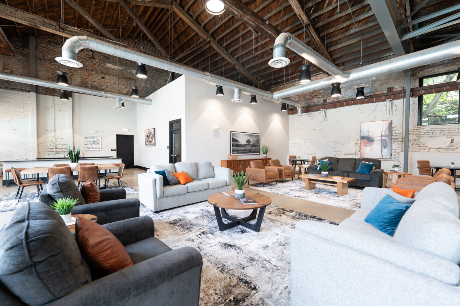 Spacious modern lounge in Columbus apartments with sofas, coffee tables, potted plants, exposed brick walls, and ceiling beams. Large windows invite plenty of natural light.