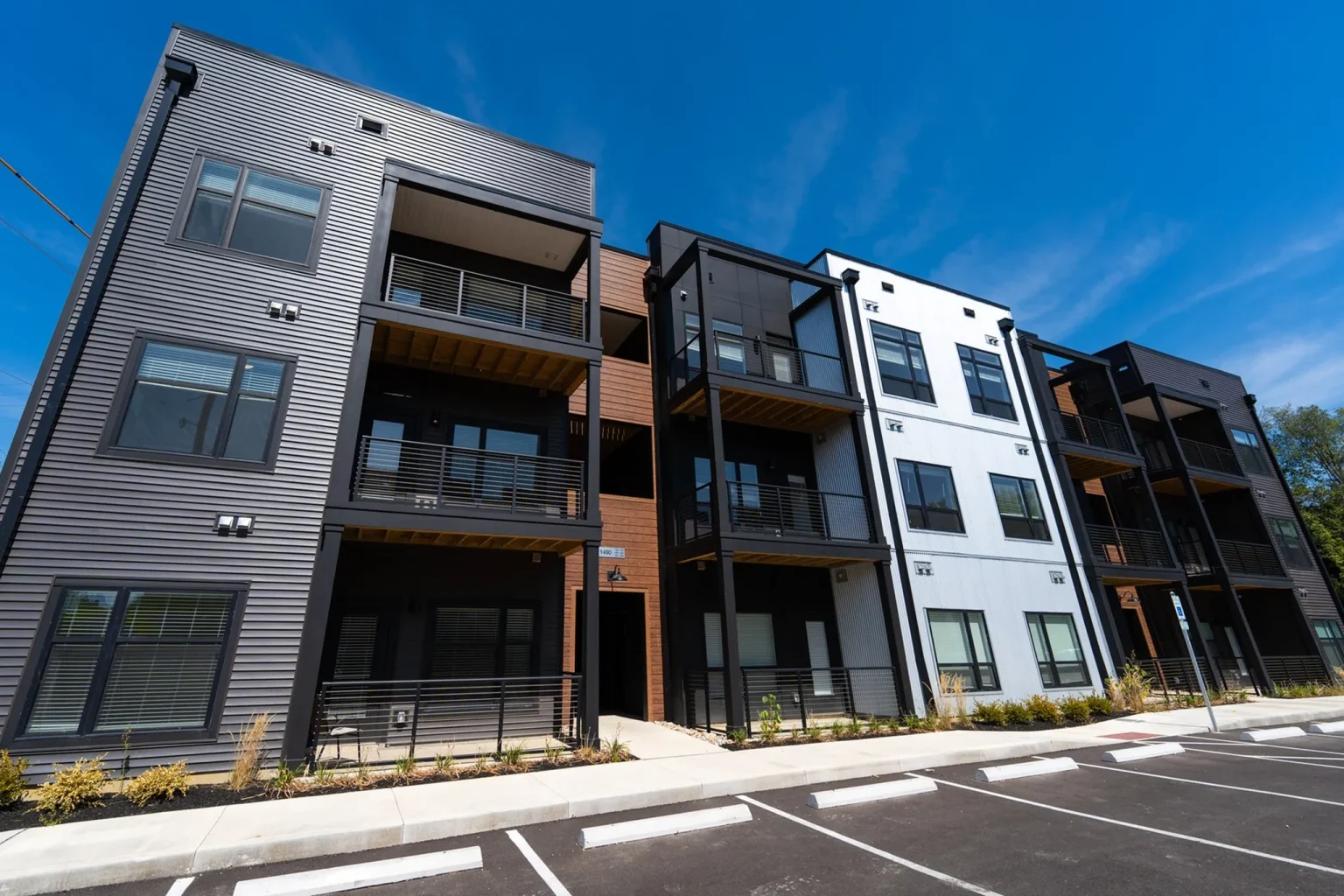 Modern three-story apartment building with a mix of black, white, and brown sections, featuring balconies and a parking lot in front.