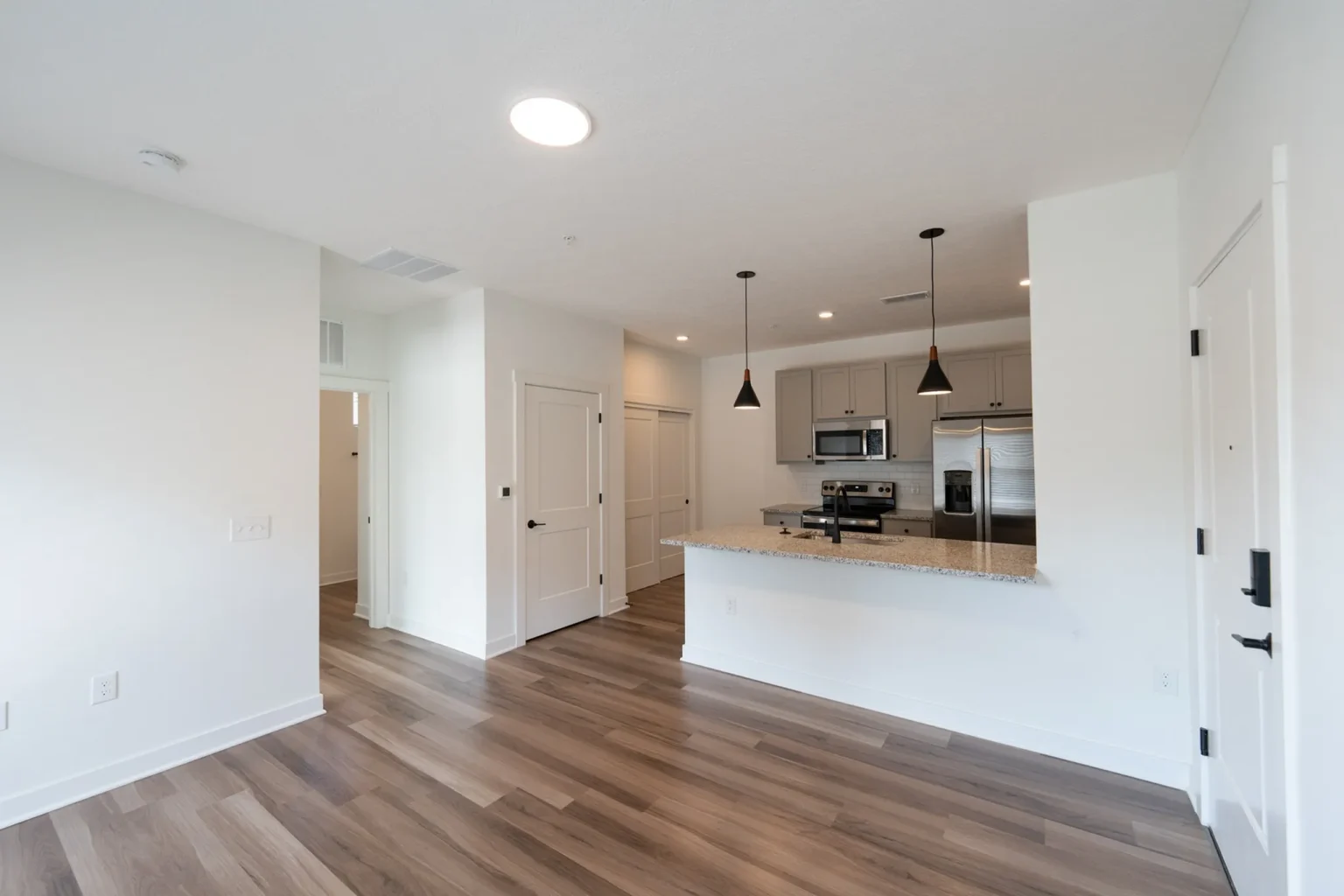 Modern kitchen with white walls, wood flooring, a granite countertop, stainless steel appliances, and pendant lighting. Open view from a living area with a round ceiling light.