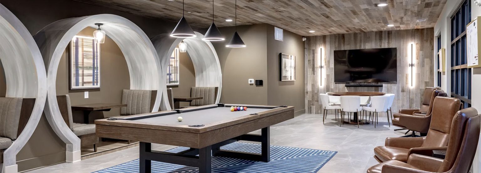 Modern game room with a pool table, lounge chairs, round table, and built-in seating. Wall-mounted TV and pendant lights complete the decor.