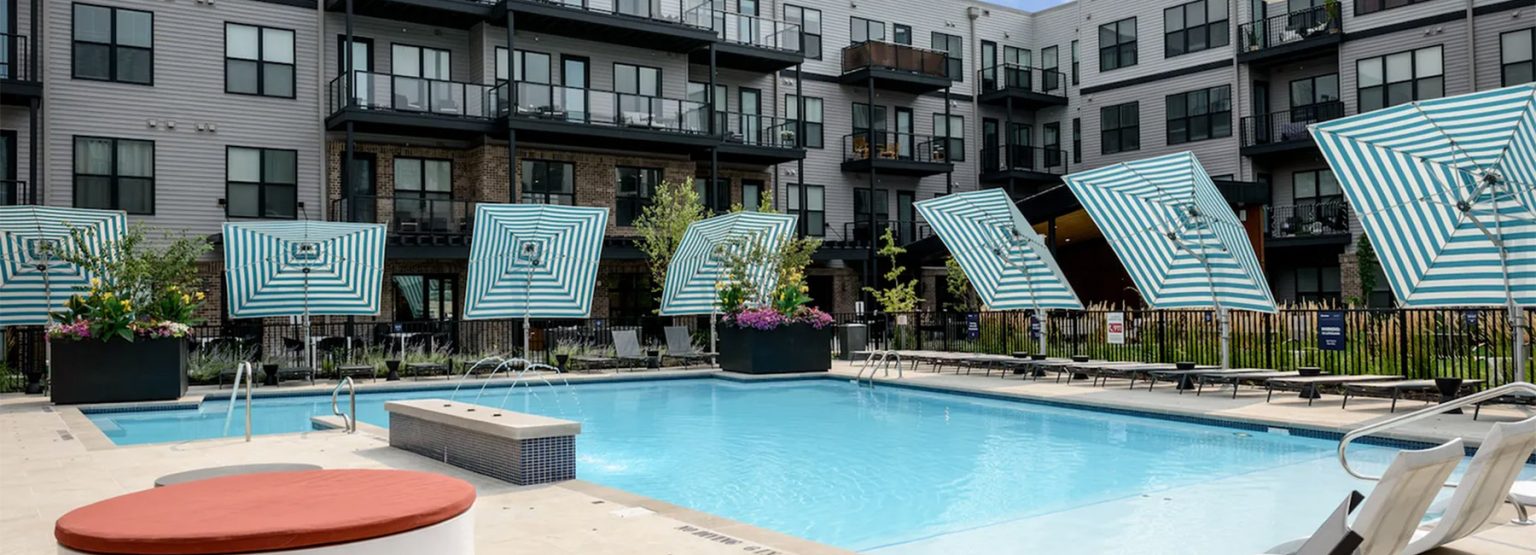 Outdoor swimming pool with blue water surrounded by lounge chairs and striped umbrellas. Modern apartment buildings with balconies are in the background.