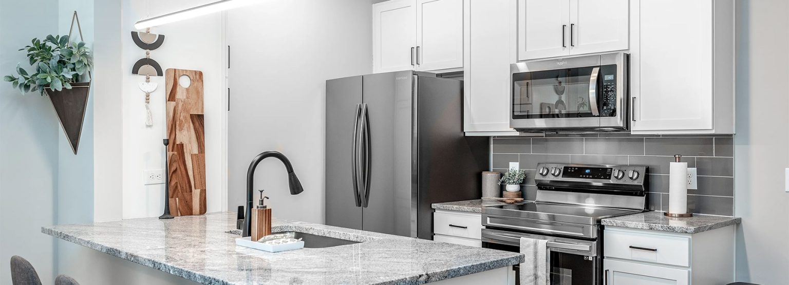 Modern kitchen with white cabinets, stainless steel appliances, gray countertops, and a textured backsplash. A potted plant and cutting boards are visible on the countertop.