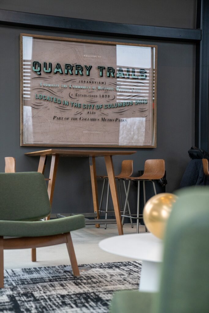 Framed Quarry Trails sign on easel in a modern room with green chairs, wooden barstools, and a patterned rug—perfect inspiration for Columbus apartments.