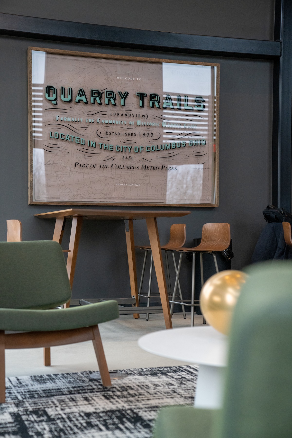 Framed Quarry Trails sign on easel in a modern room with green chairs, wooden barstools, and a patterned rug—perfect inspiration for Columbus apartments.