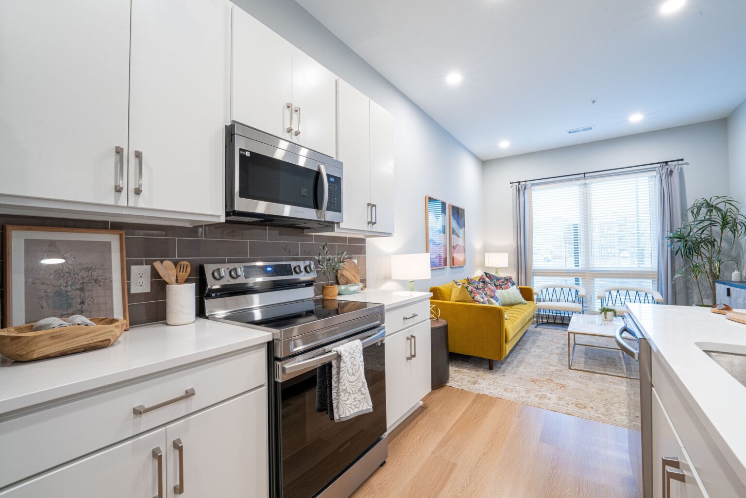 Modern kitchen with white cabinets and stainless steel appliances opens into a bright living room with a yellow sofa, large window, and light wood flooring—perfect for stylish Columbus apartments.