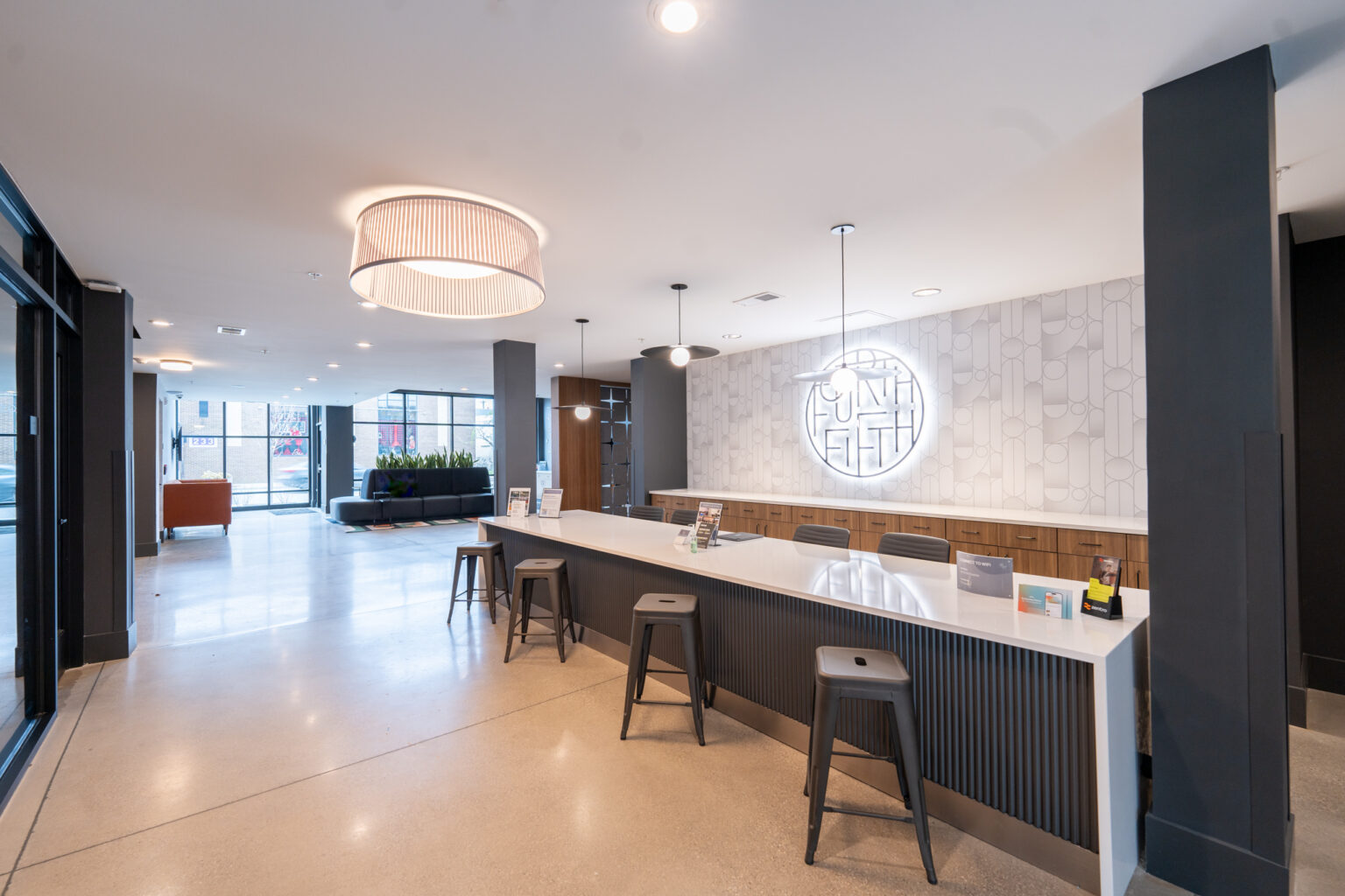 Modern lobby of Columbus apartments with a long reception desk, metal stools, minimalist decor, and a circular wall sign. Large windows provide natural light; a seating area is visible in the background.