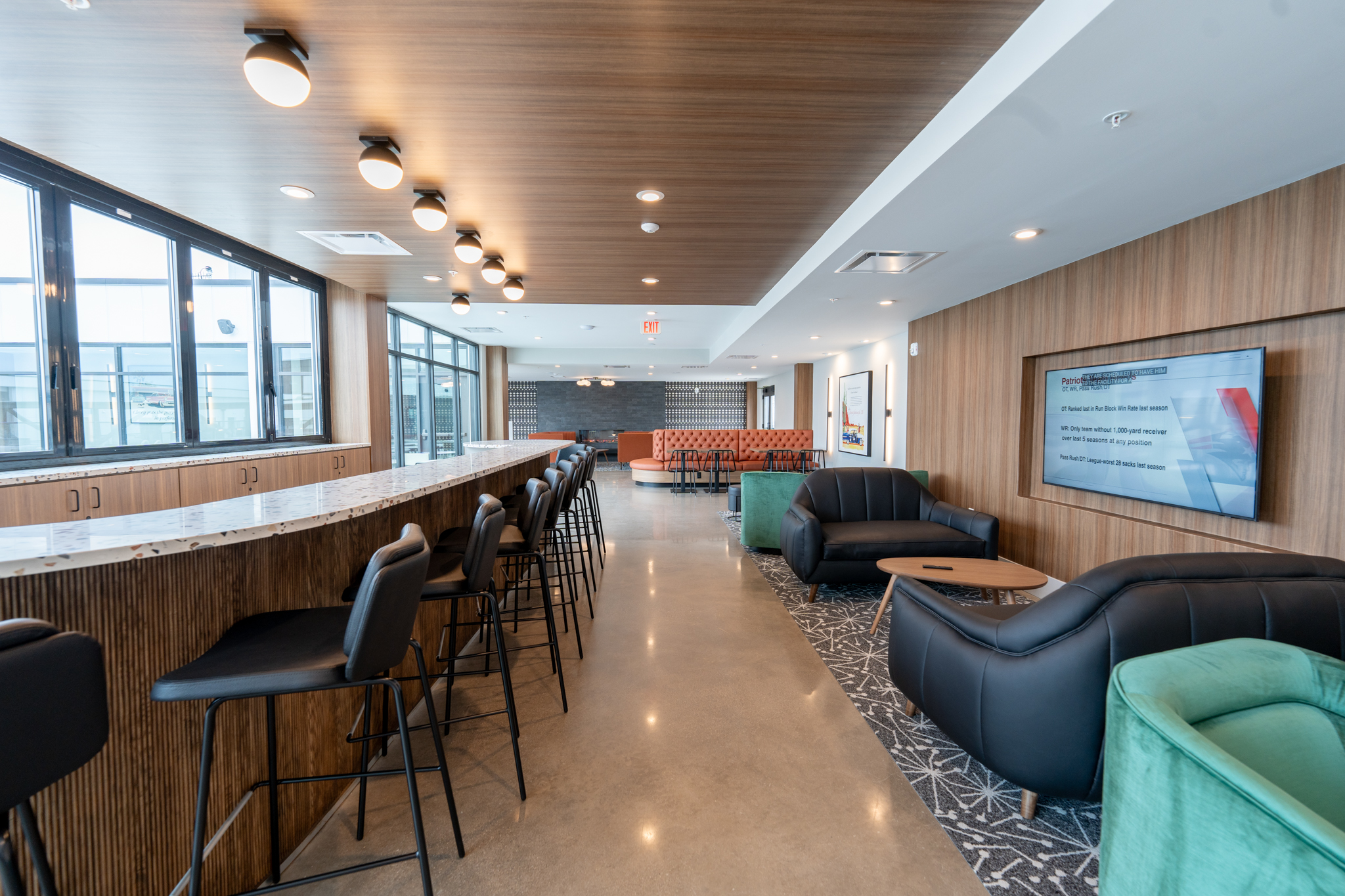 Modern lounge area in Columbus apartments with bar seating, black armchairs, a long counter, large windows, and a wall-mounted TV in a well-lit, contemporary space.