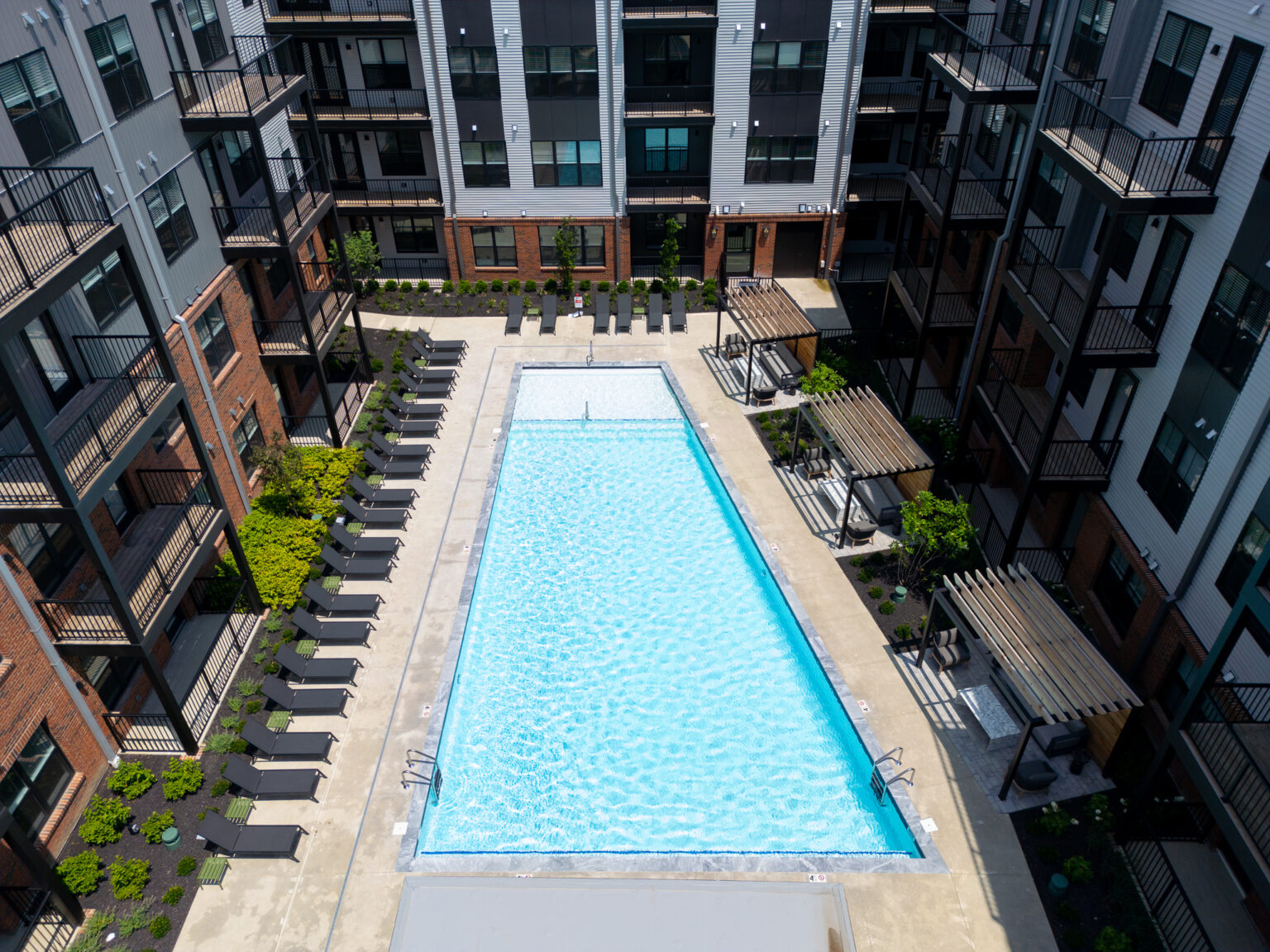 A rectangular outdoor swimming pool at Trace Quarter is surrounded by lounge chairs, lush greenery, and apartment buildings with balconies.