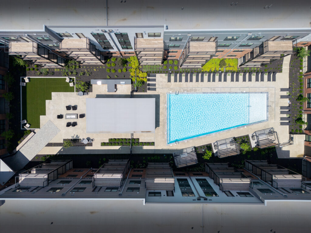 Aerial view of a rectangular outdoor swimming pool, shaded seating areas, and green spaces surrounded by modern Trace Quarter apartment buildings.