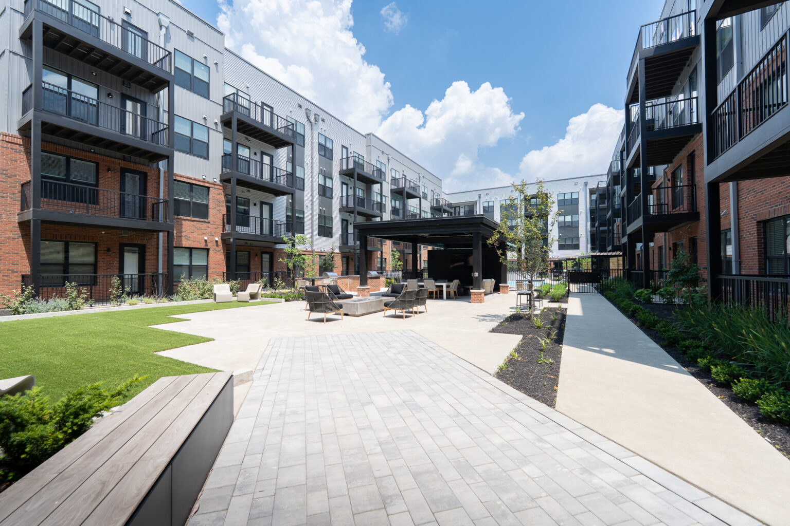 Trace Quarters modern apartment complex courtyard features inviting outdoor seating, a lush green lawn area, winding pathways, and a covered lounge space—all set beneath a partly cloudy sky.