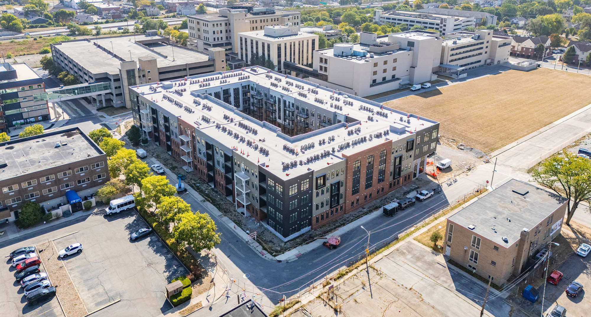 Aerial view of a large, U-shaped apartment building under construction in an urban area, surrounded by other buildings, roads, and parked vehicles.
