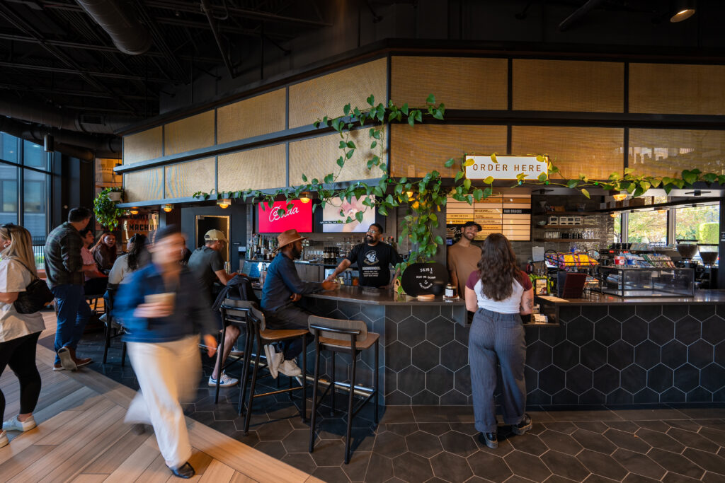 Thrive Companies, People order and interact at the busy Jeffrey Park coffee shop counter, featuring black hexagonal tiles, lush plants, and a clear Order Here sign. Apartments in Columbus Ohio