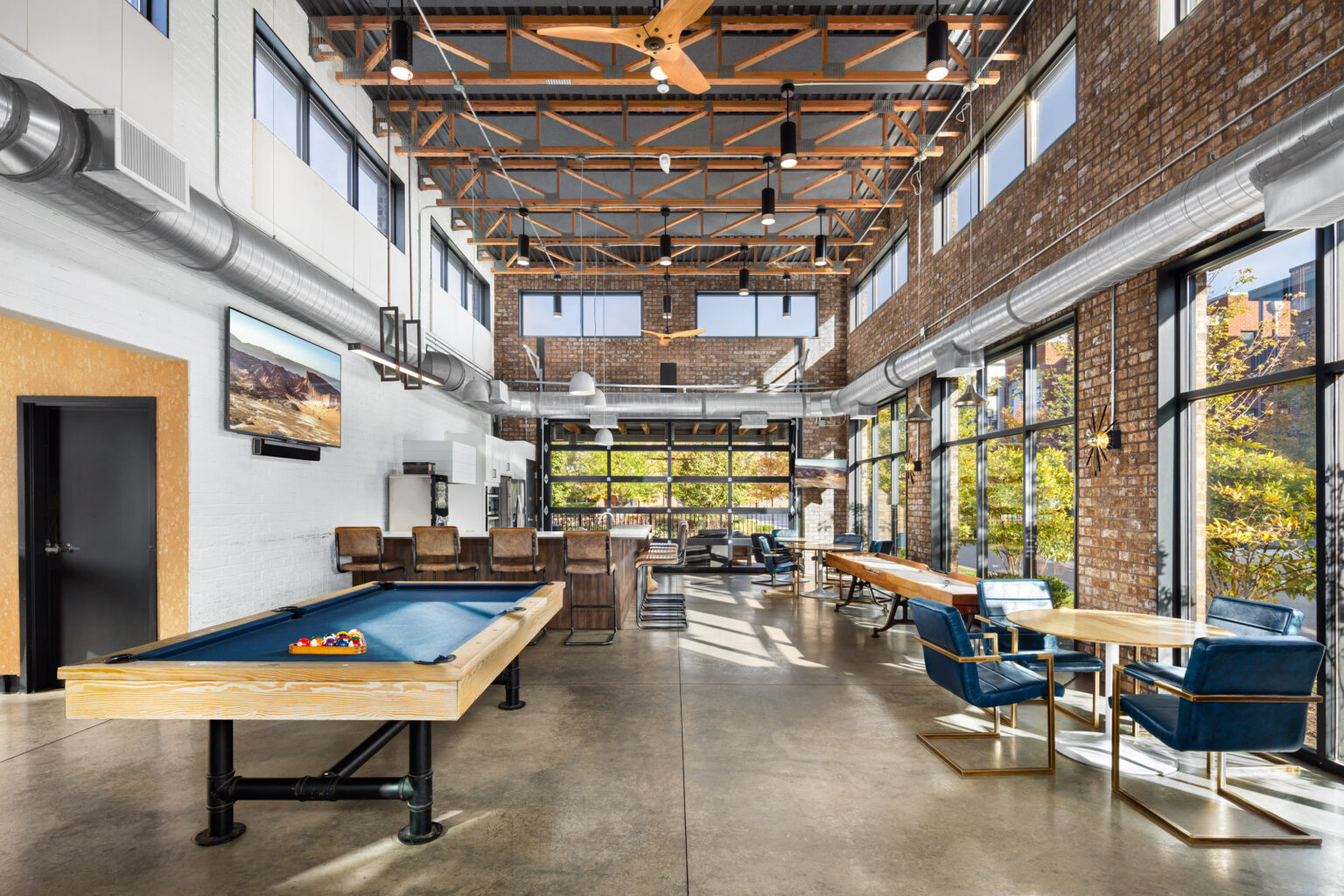 Thrive Companies, Spacious modern lounge at Jeffrey Park with a pool table, bar seating, blue chairs, large windows, exposed brick walls, and industrial ceiling beams. Apartments in Columbus Ohio