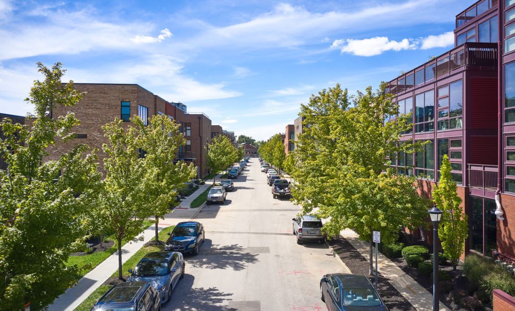 A tree-lined residential street with parked cars on both sides, flanked by modern brick buildings crafted by Thrive Companies Ohio, all under a blue sky with scattered clouds.