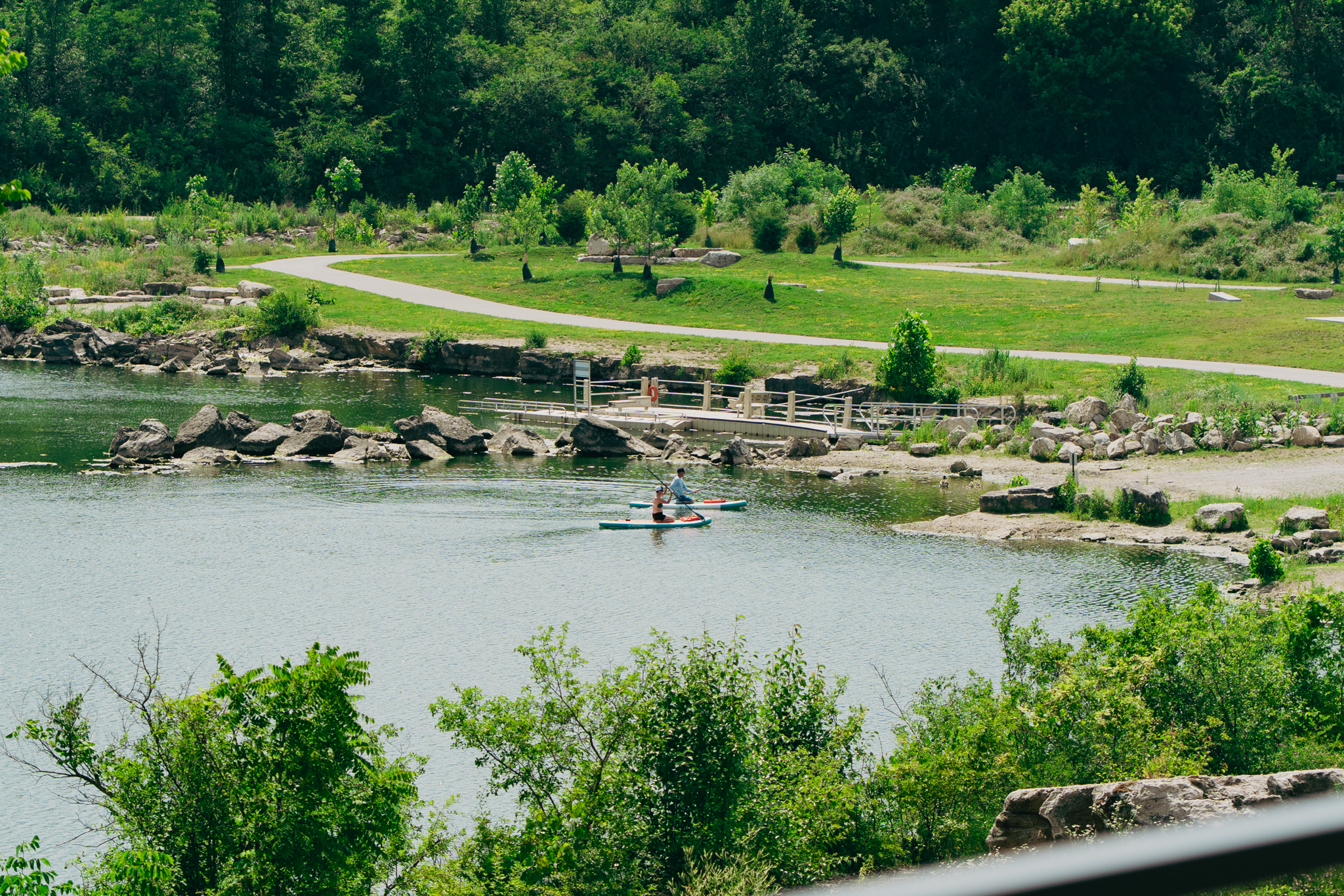 Two people paddle a canoe on a calm river at the South Bank at Quarry Trails, surrounded by greenery and rocks, with a trail and footbridge visible in the background.